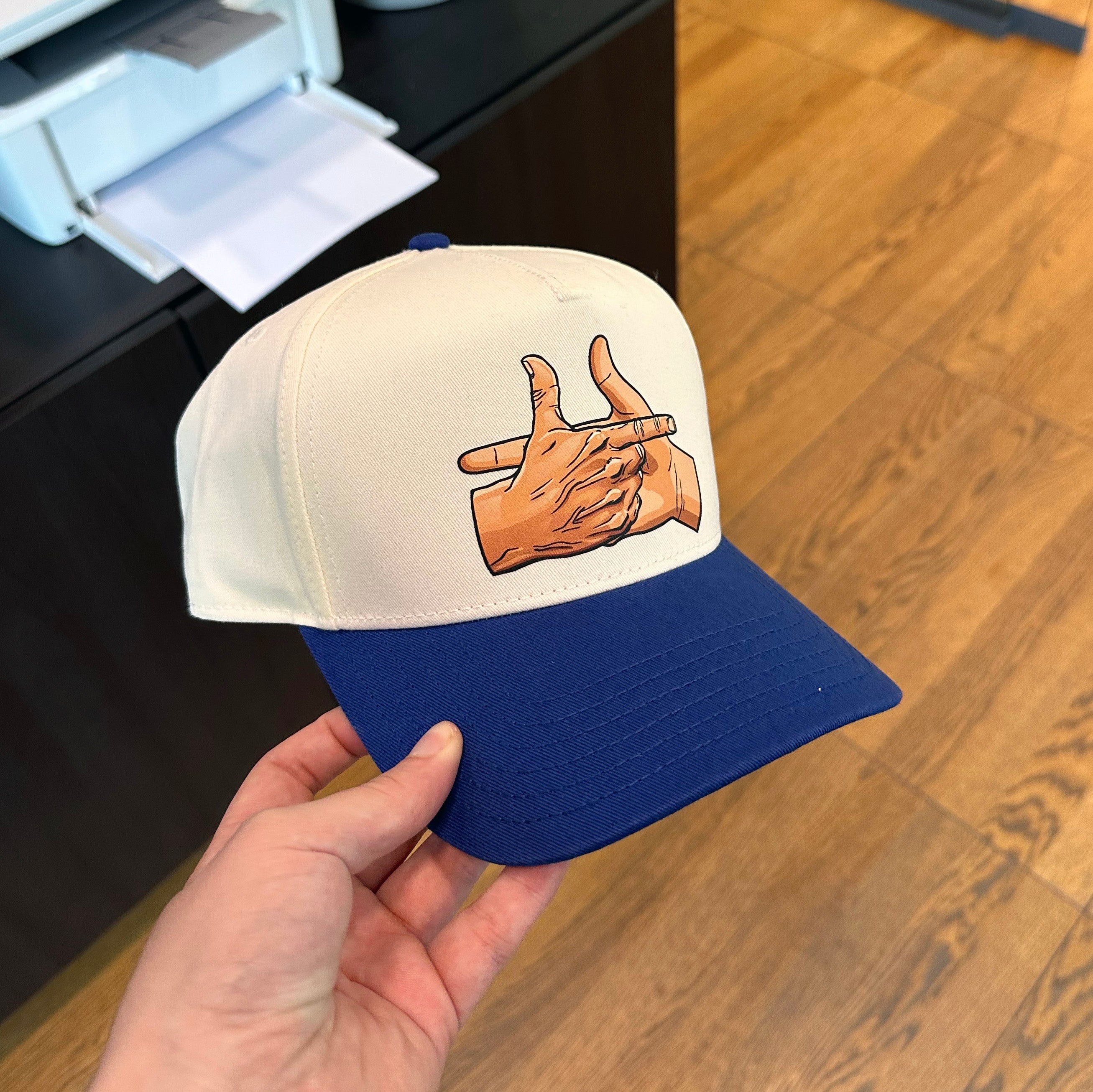 Cap with hand gesture design held by a hand on a wooden floor.