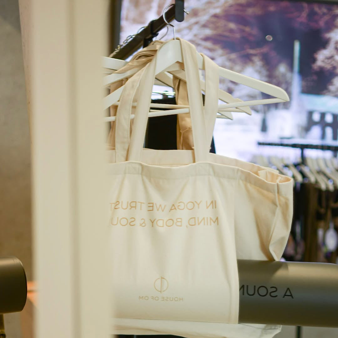 Beige tote bag with gold text on a hanger against a blurred indoor background